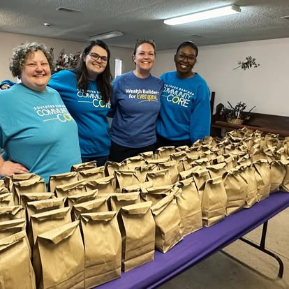 Four volunteers in Community Core shirts stand behind a table covered in packed paper lunch bags